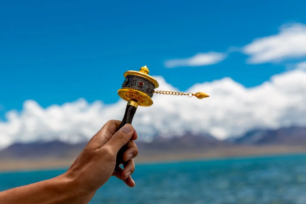 Hand holding a Tibetan prayer wheel with a serene lake, white clouds, and majestic mountains in the background.